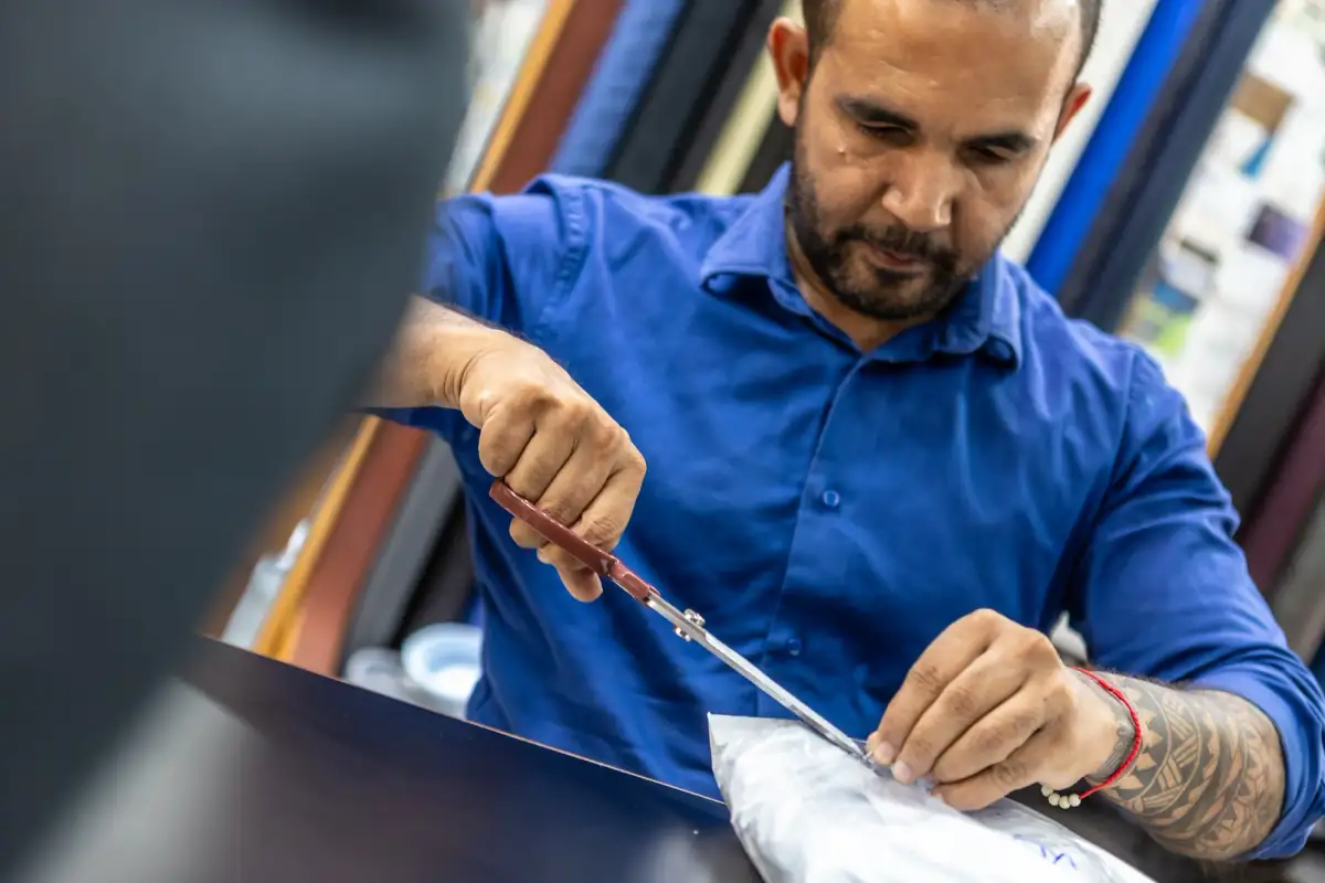 Tailor in a blue shirt cutting shirting cloth with shears at the workbench in Patong